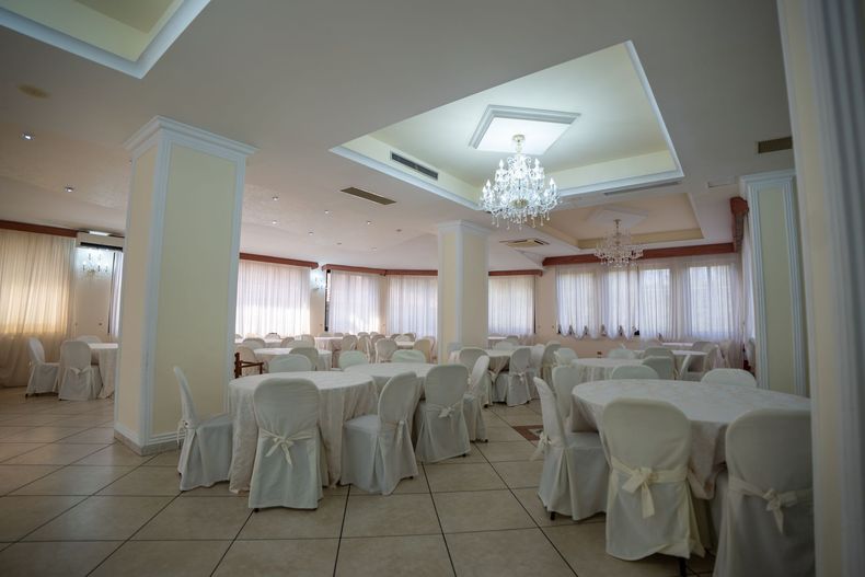 Sala da pranzo con tavoli e sedie drappeggiati di bianco, lampadari e finestre con tende bianche.