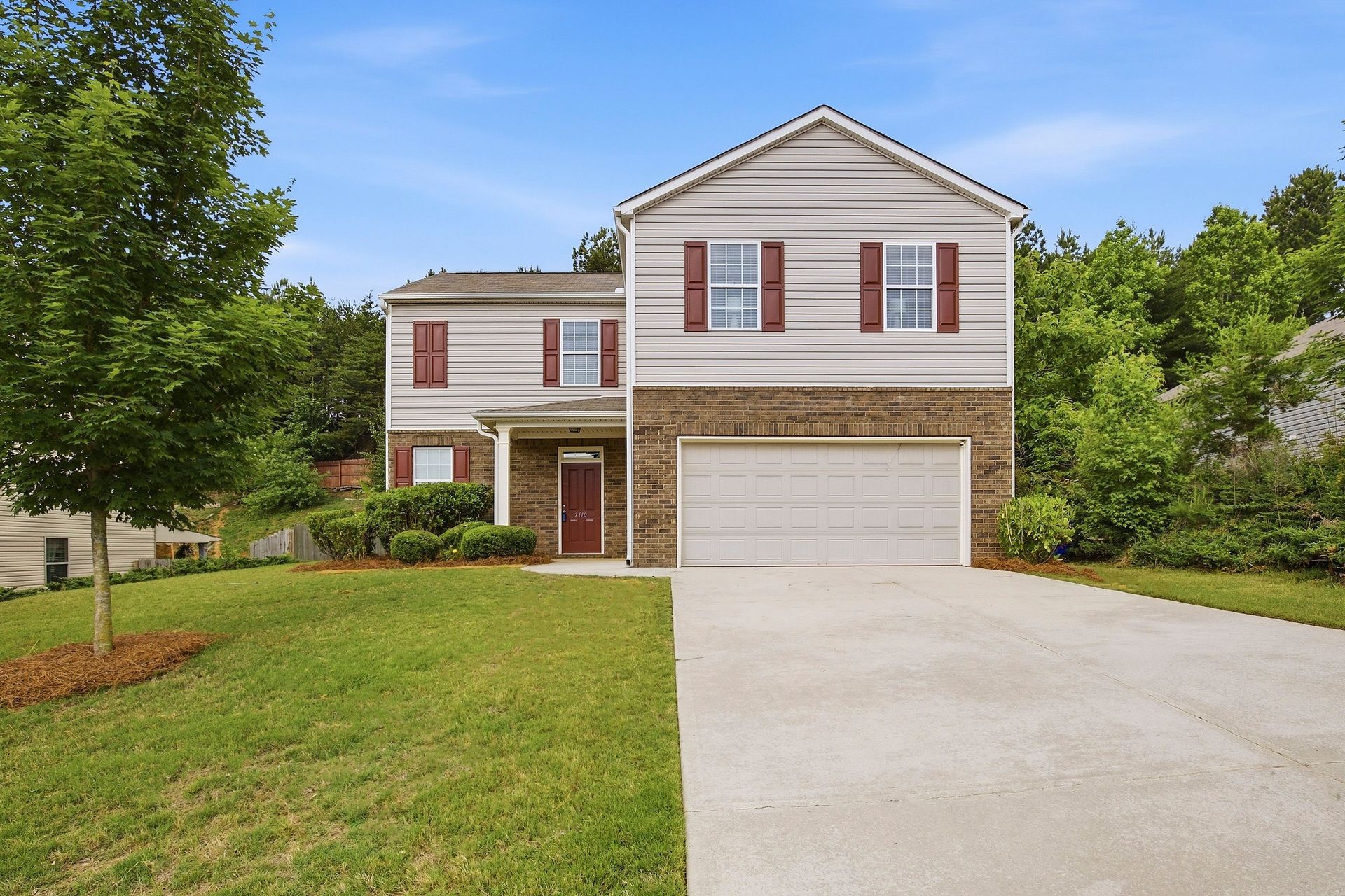 Suburban two-story house with beige siding, brick accents, attached garage, and a long driveway.