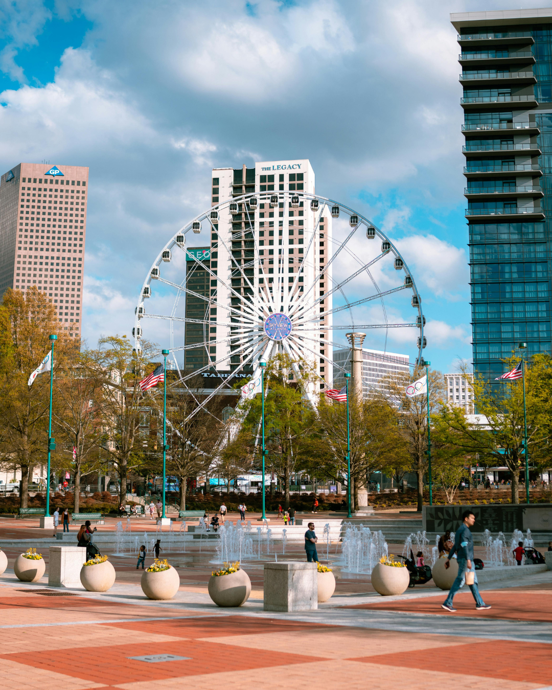 Ferris wheel in a park with fountains, trees, and buildings against a cloudy blue sky. People walk around.