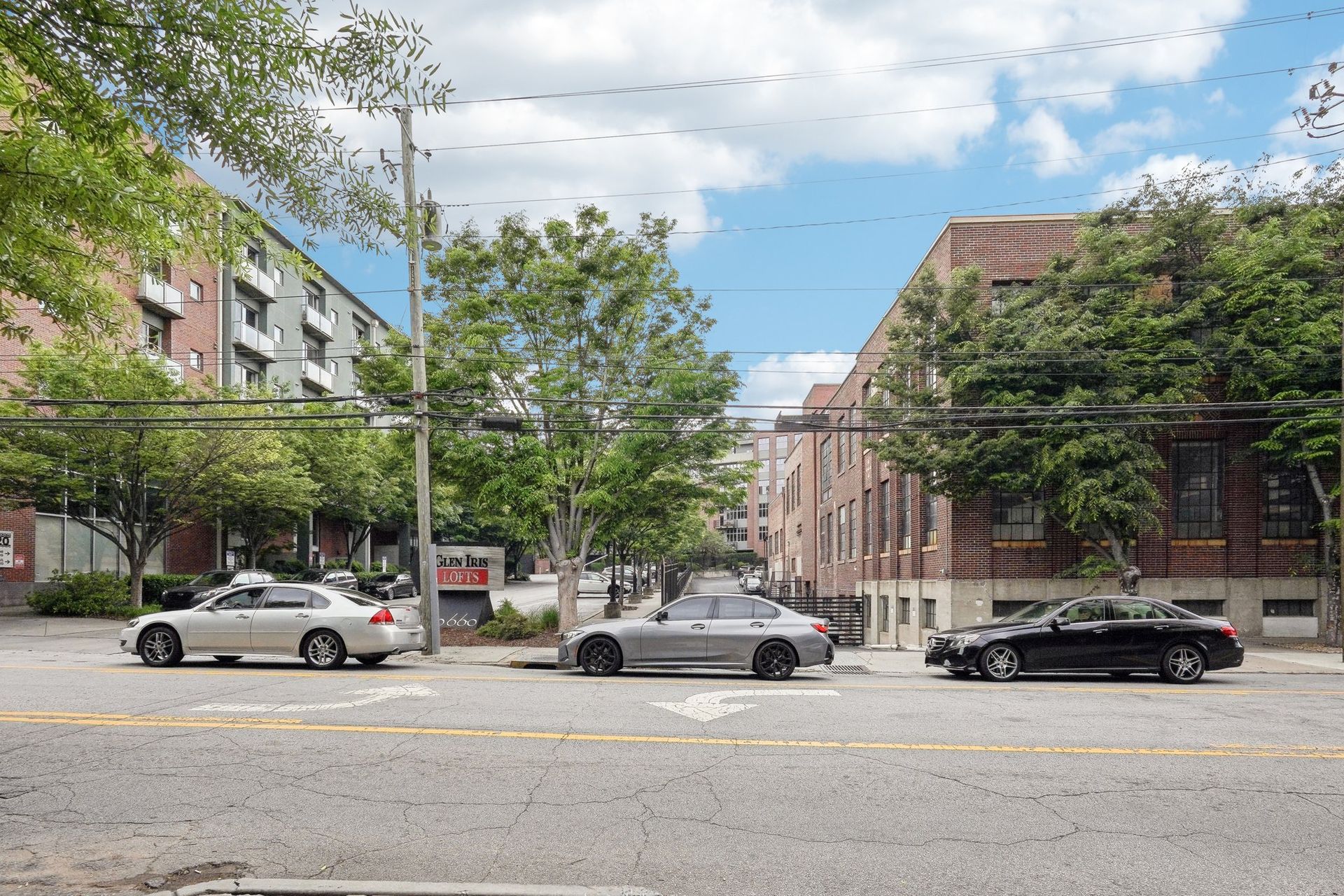 Cars parked on a street lined with buildings and trees, under a partly cloudy sky.