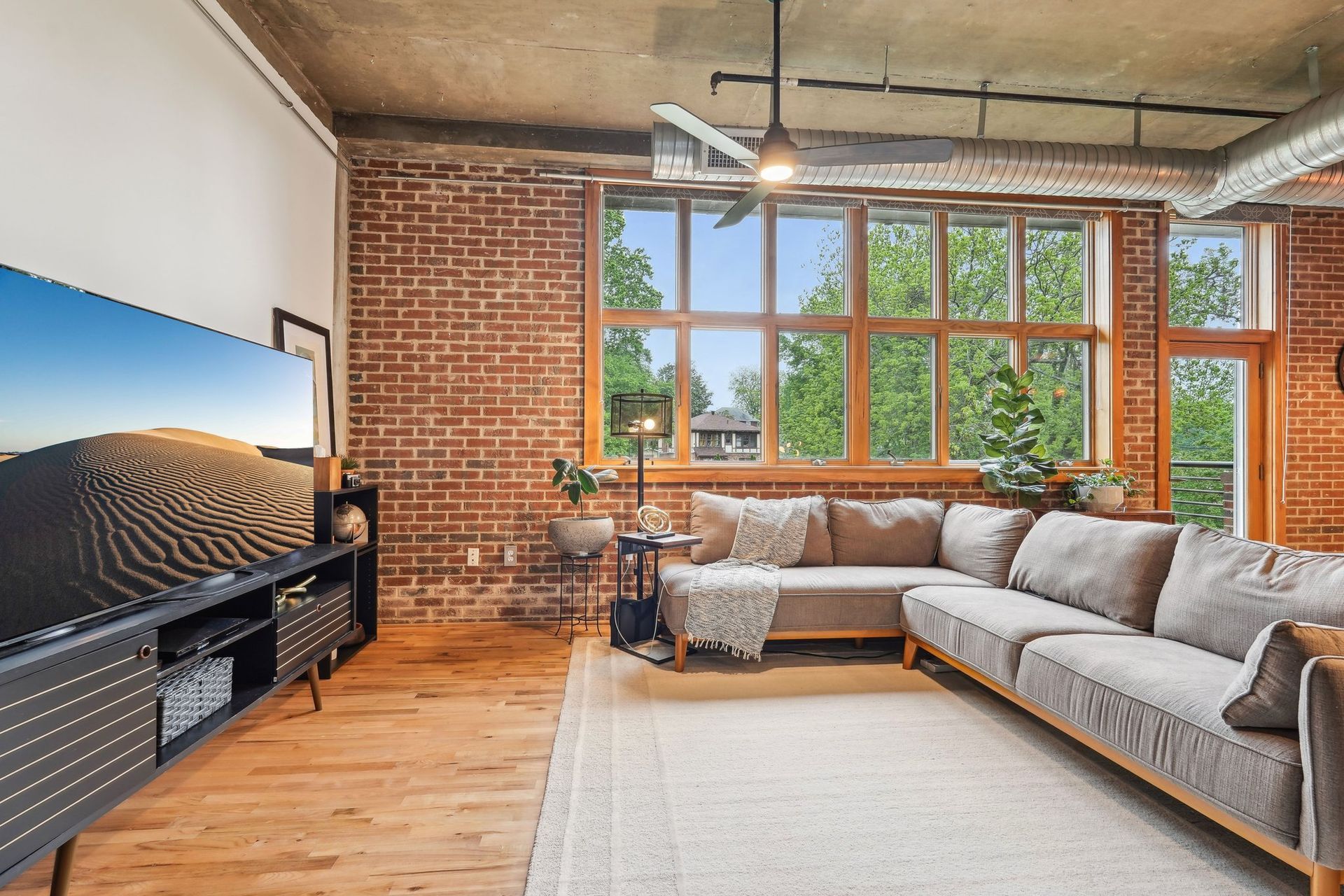 Living room with exposed brick wall, large windows, L-shaped sofa, and TV.
