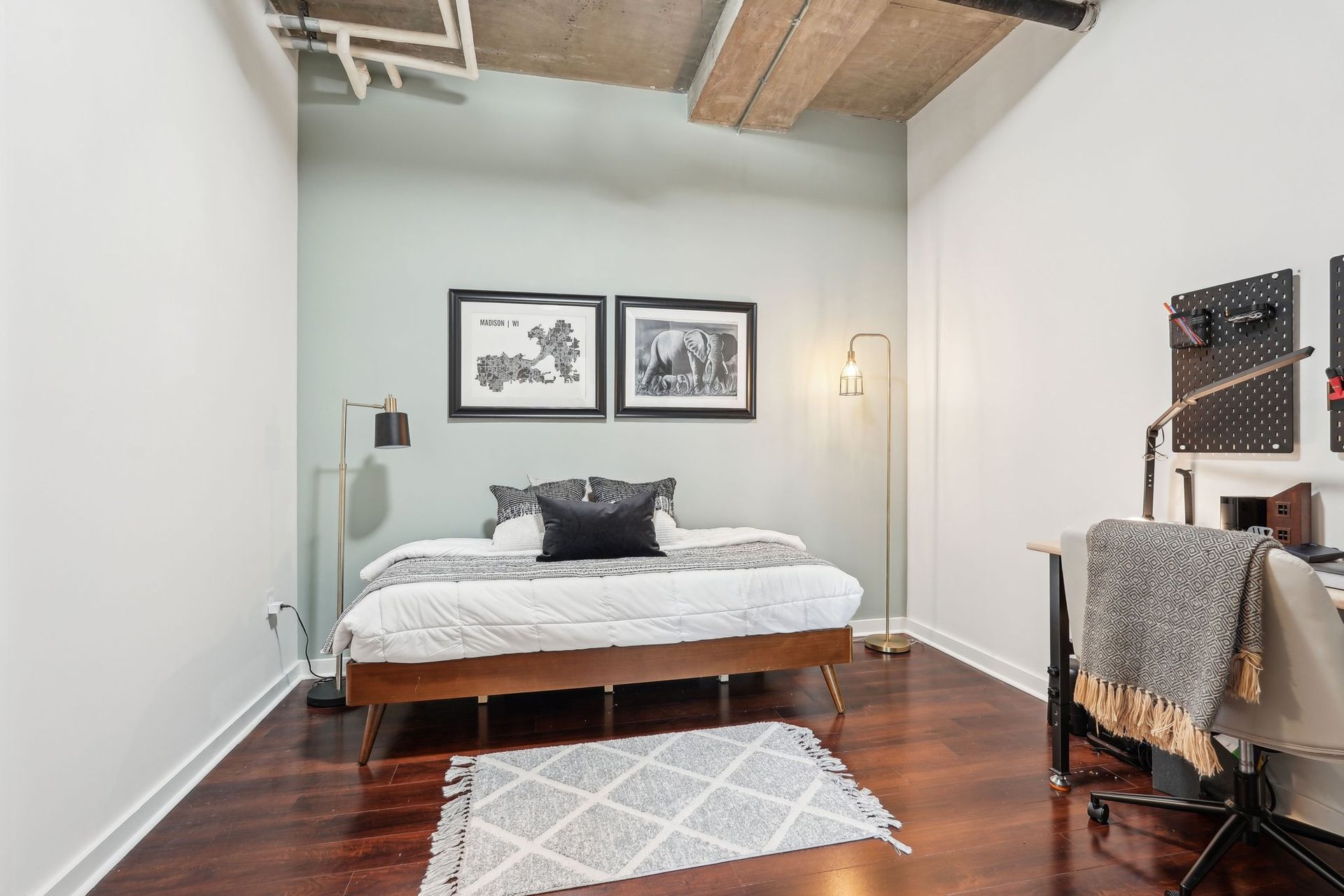 Bedroom with bed, desk, artwork, and area rug. Walls are white and green, with wood flooring.