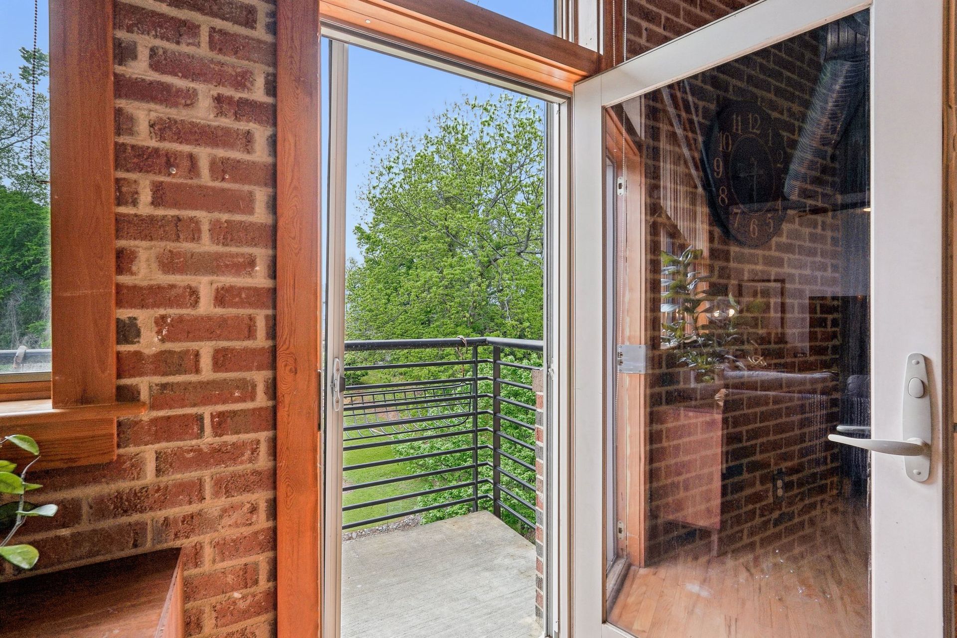 View of a small balcony with a metal railing, framed by glass doors and a brick wall; trees in the background.