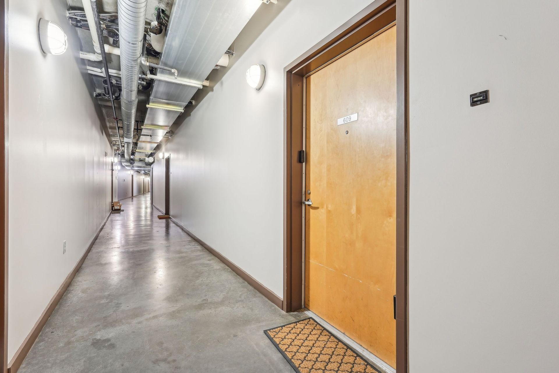 Long hallway with a closed wooden door, concrete floor, and overhead pipes.