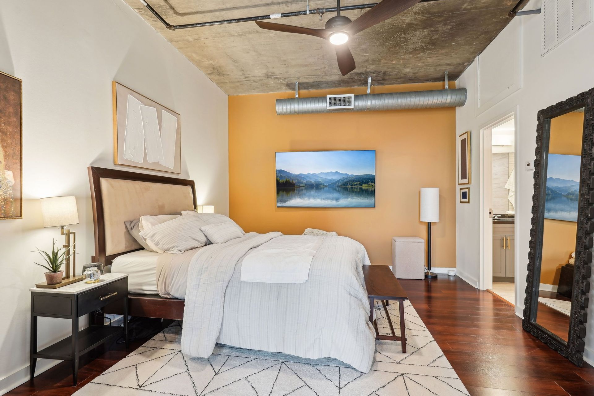 Bedroom with exposed ceiling, bed with white bedding, TV on mustard-colored wall, full-length mirror, wood floors.