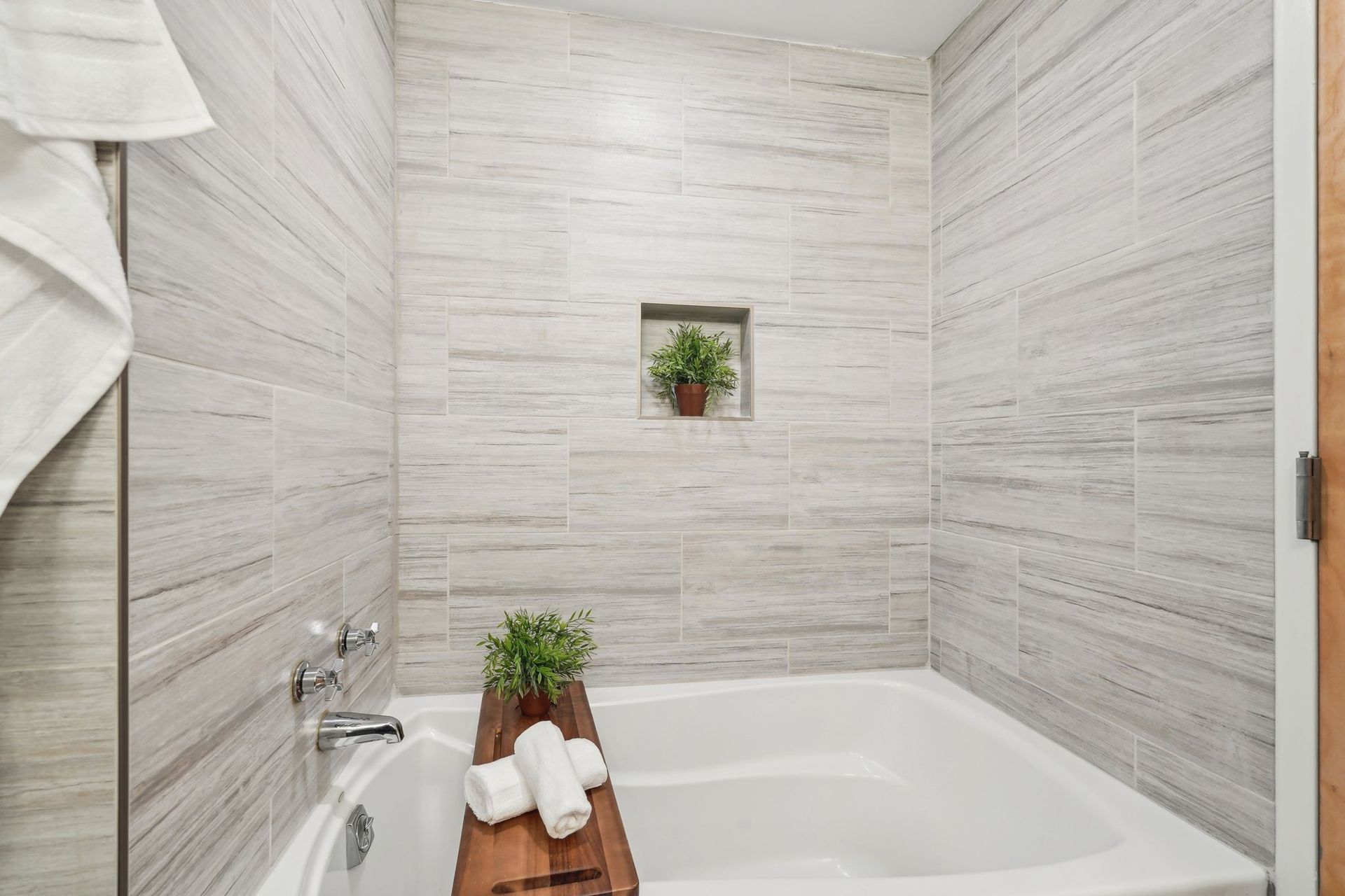 Bathroom with light-colored, patterned tile, a white bathtub, and a built-in shelf with a plant.