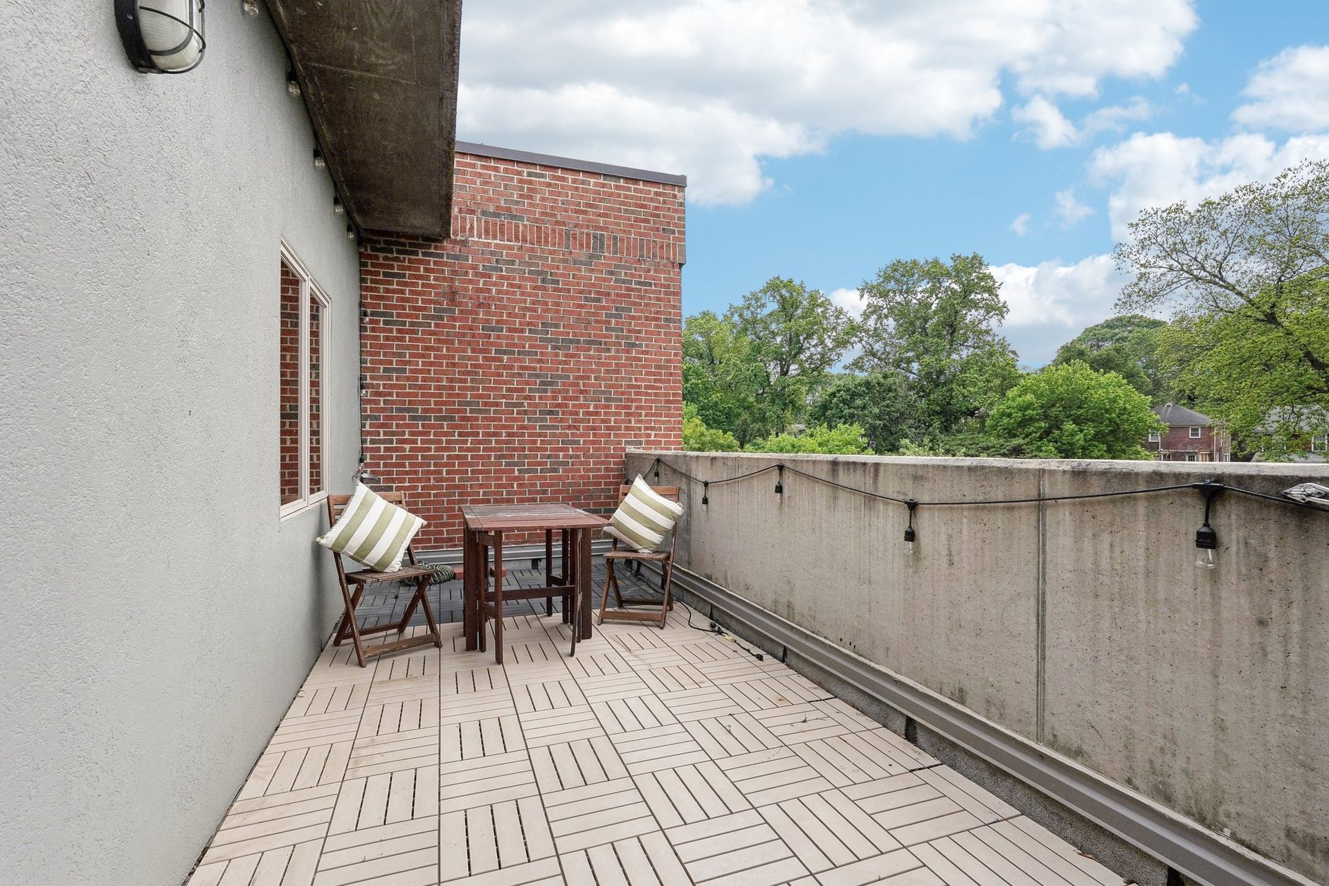 Small outdoor balcony with a table and chairs, brick wall, and trees in the background under a cloudy sky.