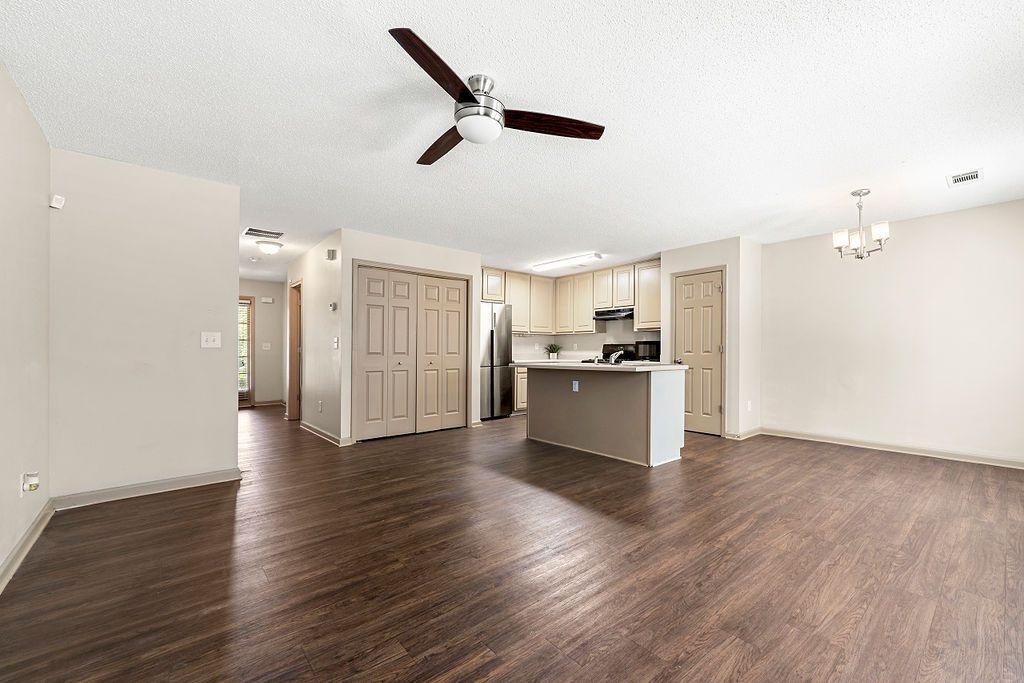 An empty living room with a ceiling fan and a kitchen in the background.