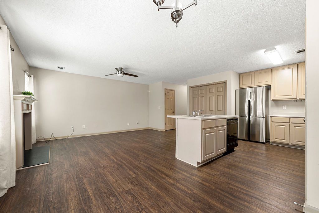 An empty living room with hardwood floors and a kitchen with stainless steel appliances.