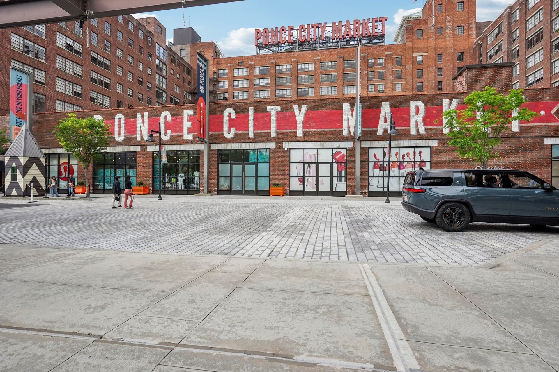 Exterior view of Ponce City Market building with shops and parked car. Red brick, gray cobblestone.