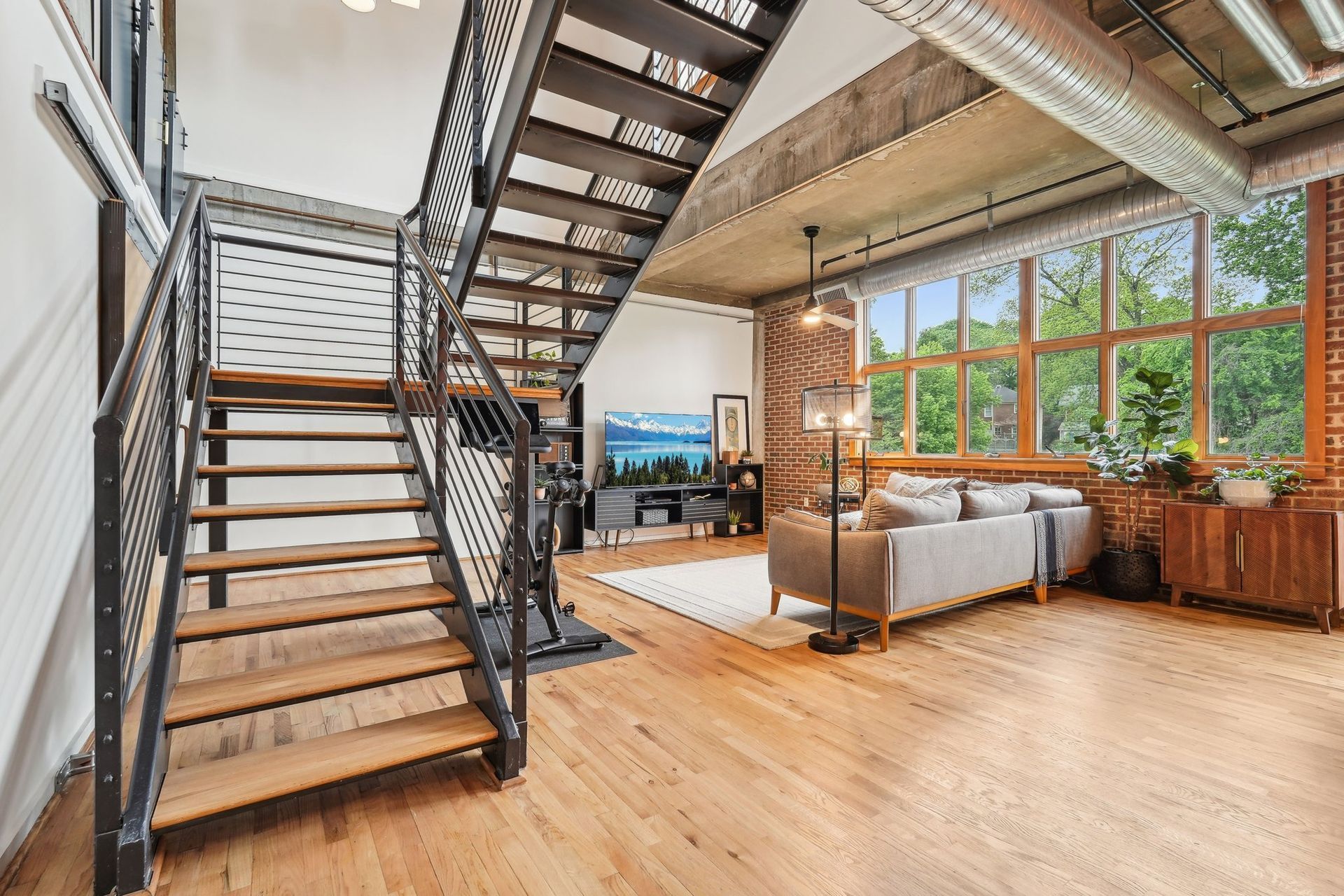 Two-story loft with metal staircase, hardwood floors, large windows overlooking trees, and a gray sofa.