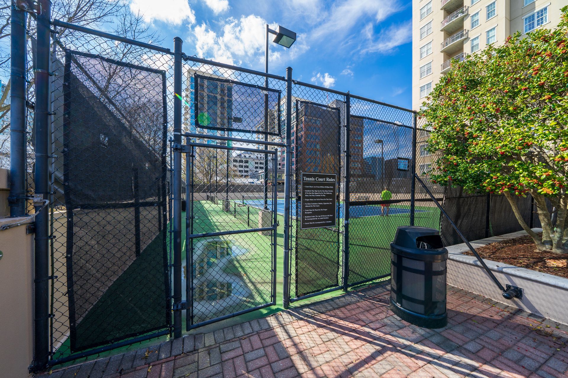 A tennis court is behind a chain link fence with a trash can in front of it.