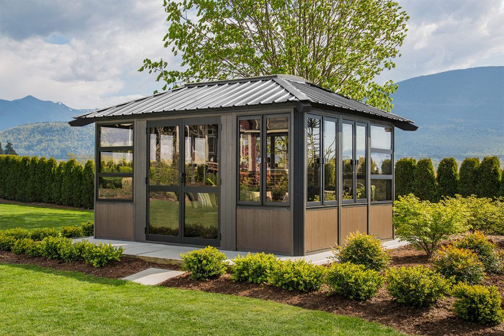 Gazebo with large windows, brown siding, gray roof, on a green lawn with a mountain backdrop.