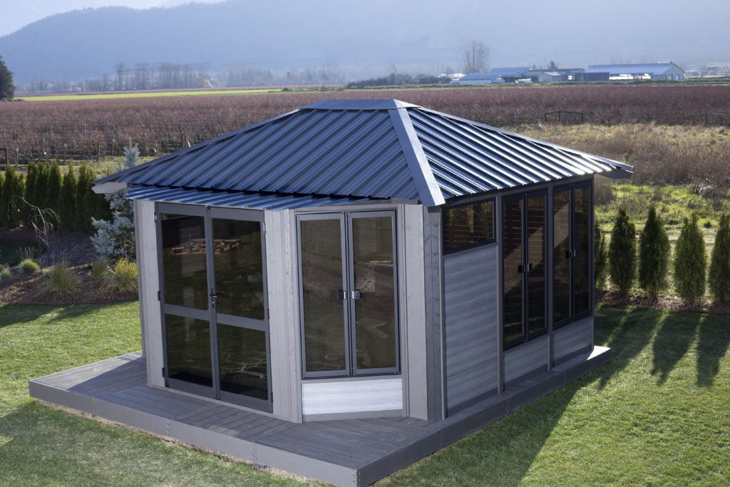 Gray gazebo with dark roof, windows, and surrounding green lawn against a field and mountain backdrop.