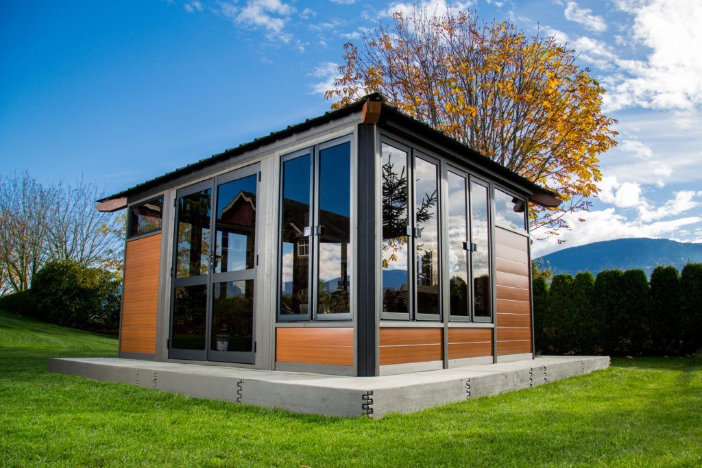 Gazebo with glass windows and brown siding on a concrete base, set in a grassy yard under a blue sky.