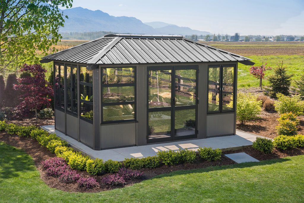 Gazebo with dark frame and windows on a concrete patio, surrounded by landscaping.