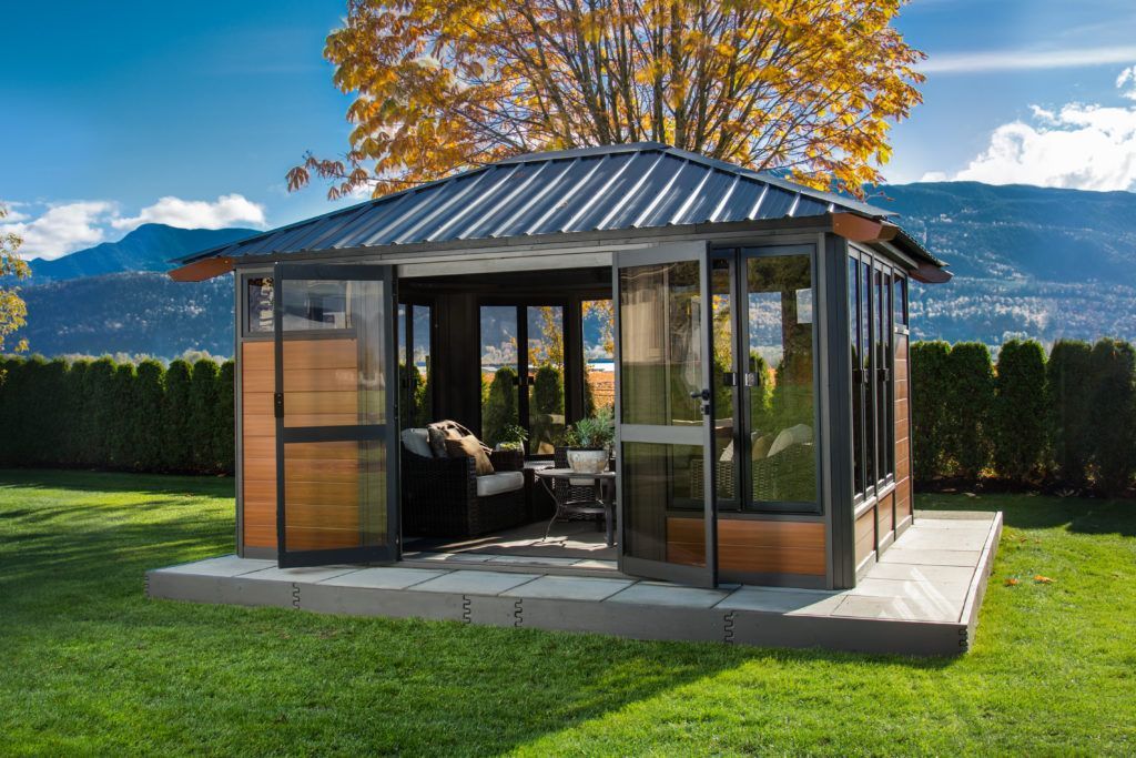 Gazebo with glass walls, metal roof, wooden accents, on a stone base, green lawn, mountains in the background.
