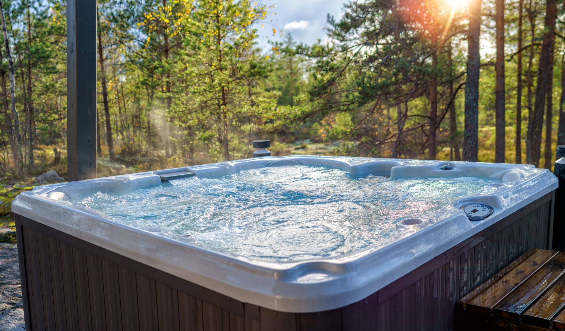 A bubbling hot tub on a deck overlooking a forest at sunset, with sunlight filtering through the trees.
