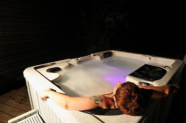 Person relaxing in outdoor hot tub at night with purple lights and steam rising from warm water.