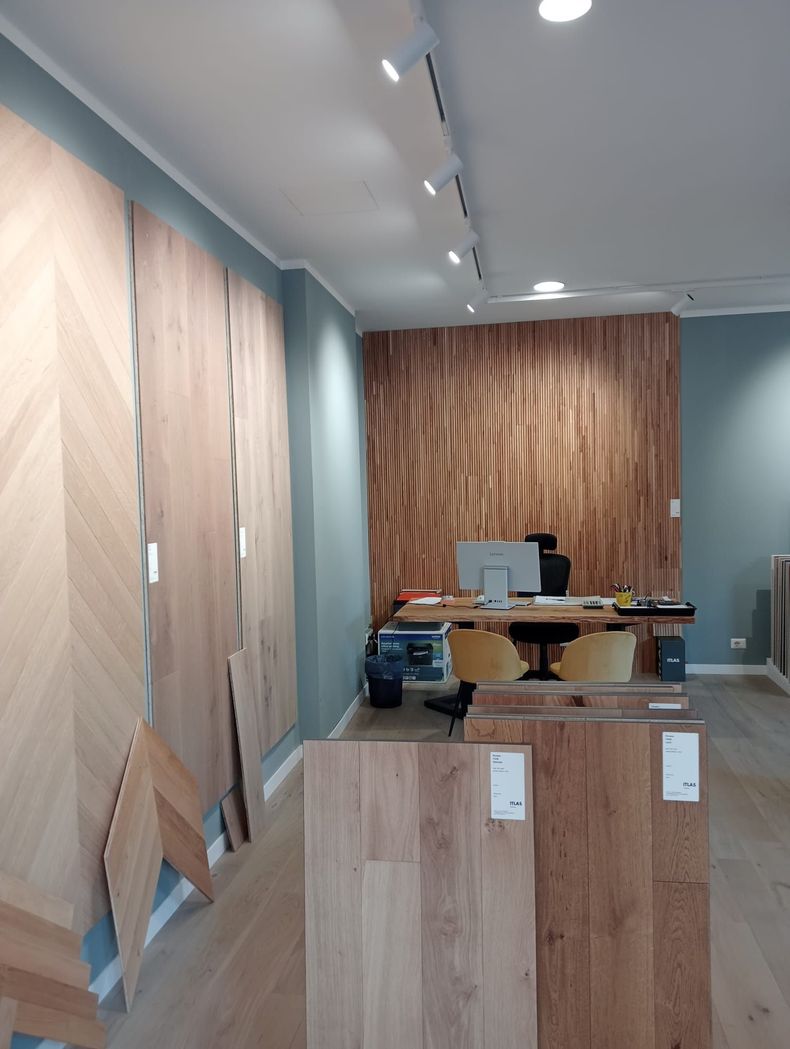 Showroom con vari campioni di pavimenti in legno e zona scrivania con computer. Pannelli in legno chiaro alle pareti.