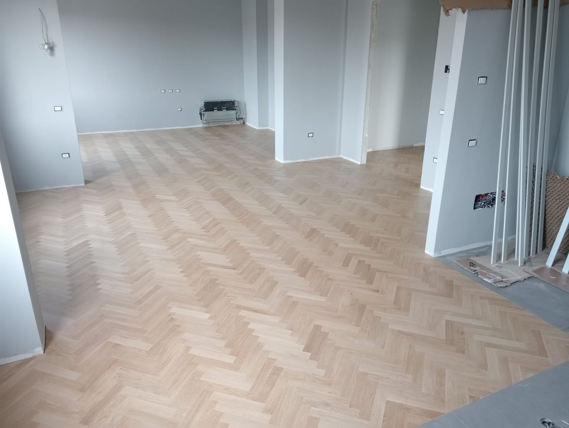 Pavimento in legno chiaro a spina di pesce in una stanza con pareti grigio chiaro e finiture bianche.