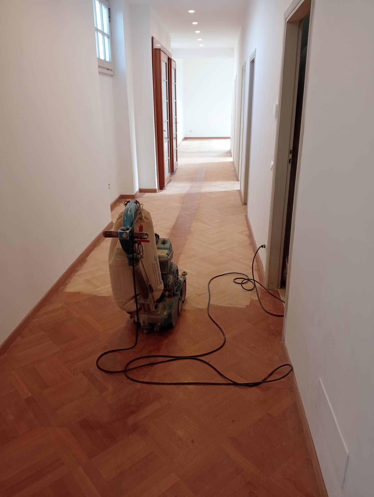 Un lungo corridoio con un pavimento in legno in fase di levigatura. In primo piano una levigatrice.