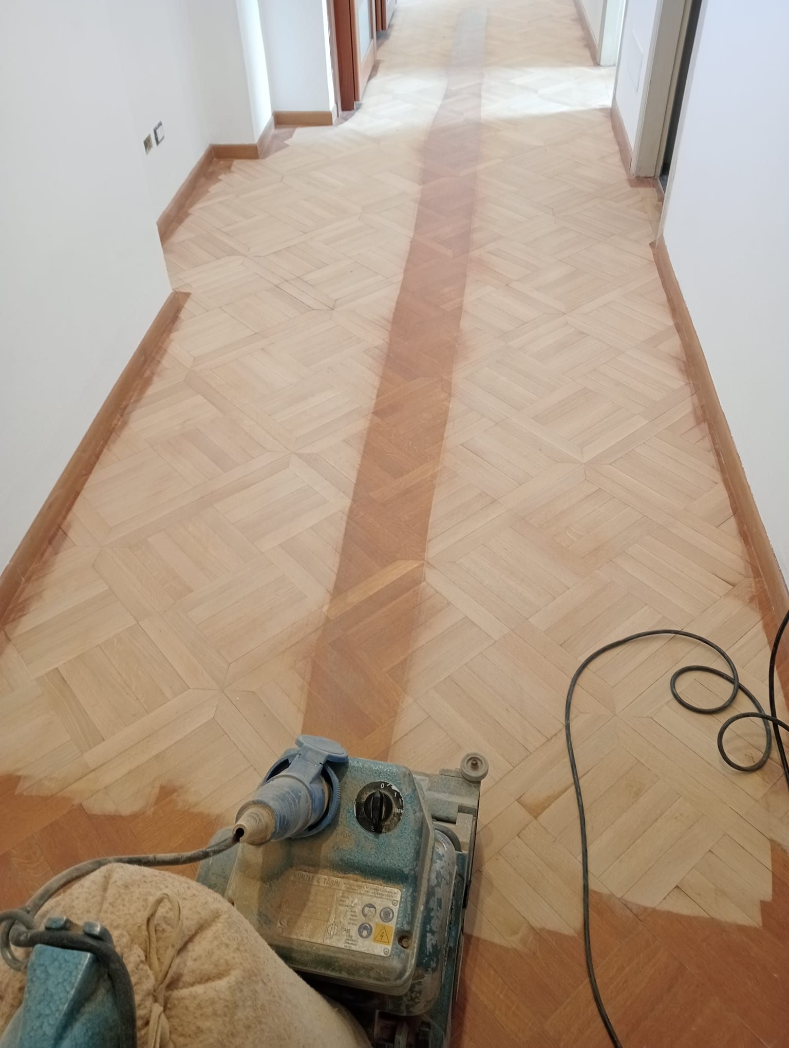 Un corridoio con pavimento in parquet in fase di levigatura. Una levigatrice viene utilizzata attivamente su una striscia di legno centrale. Pavimento beige chiaro e pareti bianche.