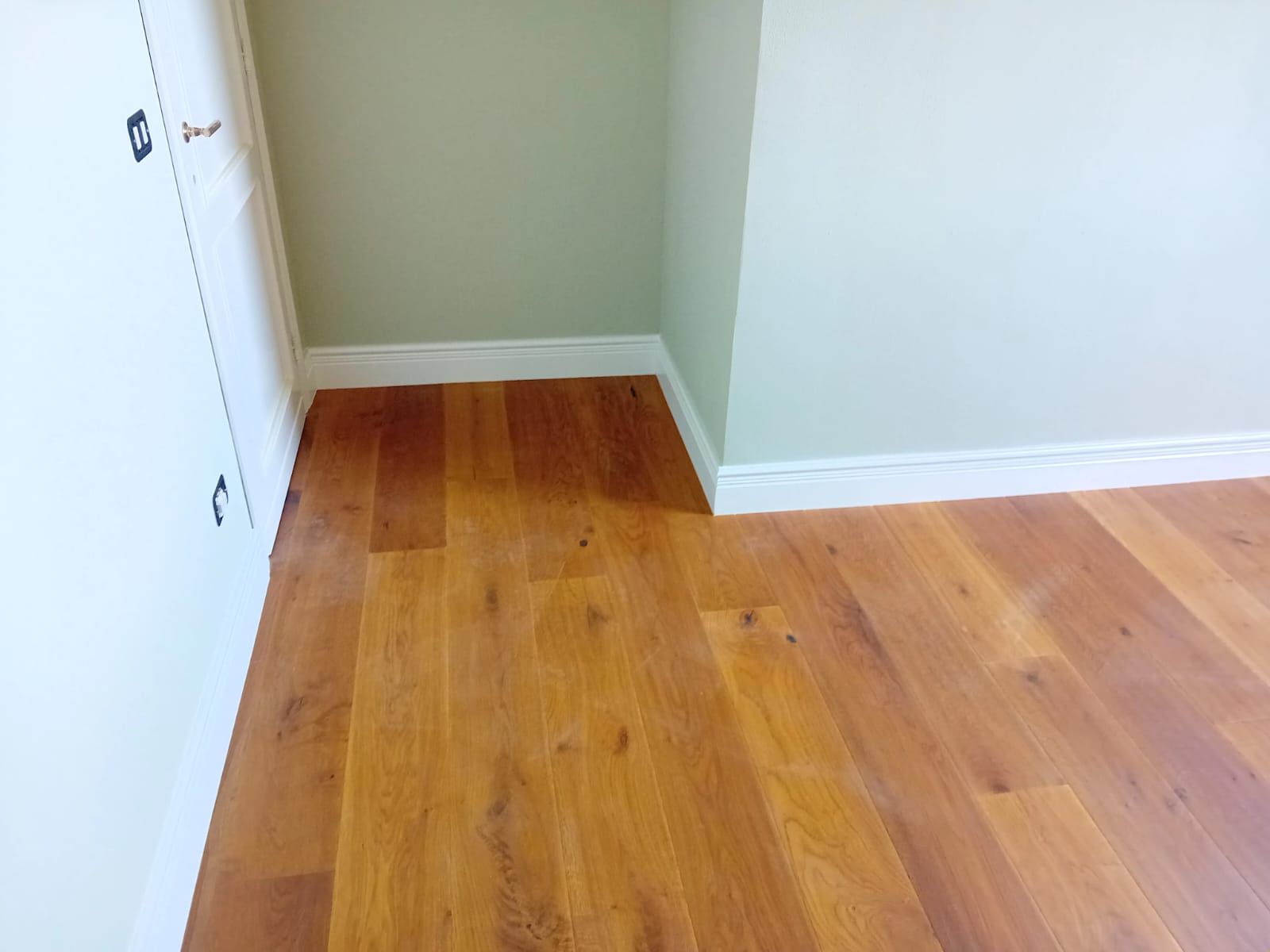 Pavimento in legno in una stanza con pareti bianche e verde chiaro, battiscopa bianchi e stipite della porta bianco.