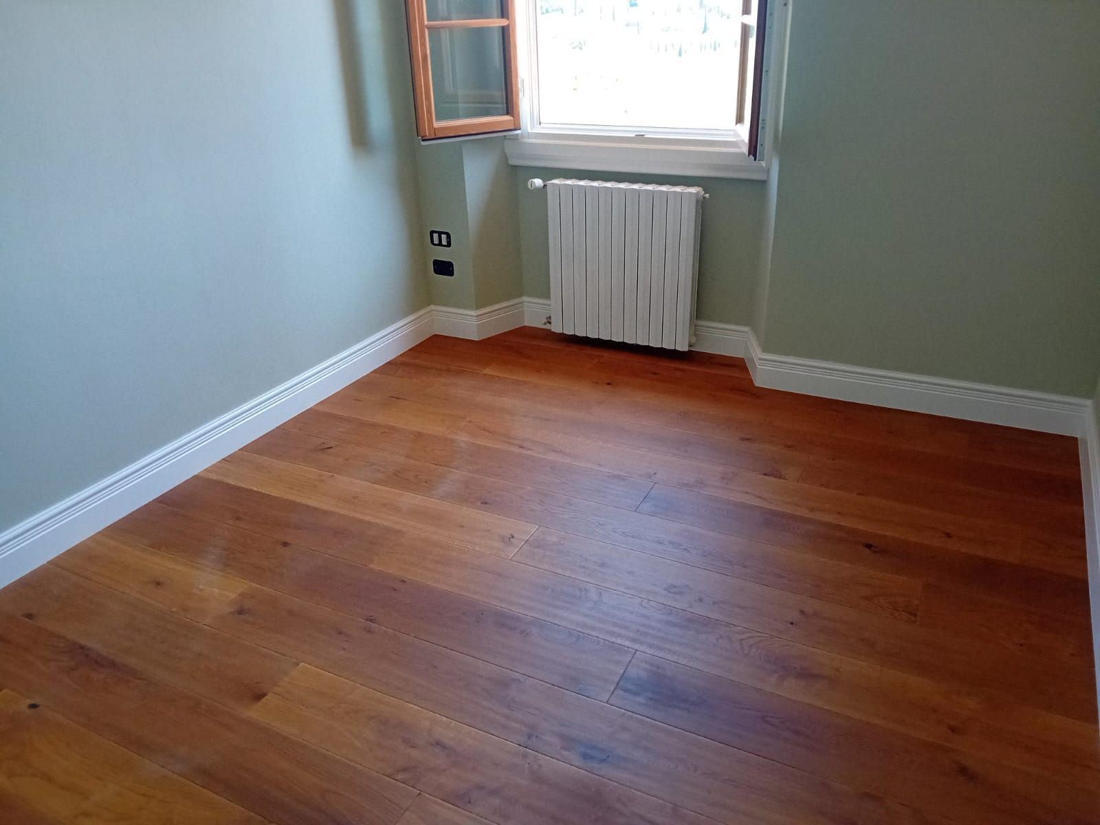 Stanza vuota con pavimento in legno, pareti verde chiaro, finiture bianche, termosifone sotto una finestra.