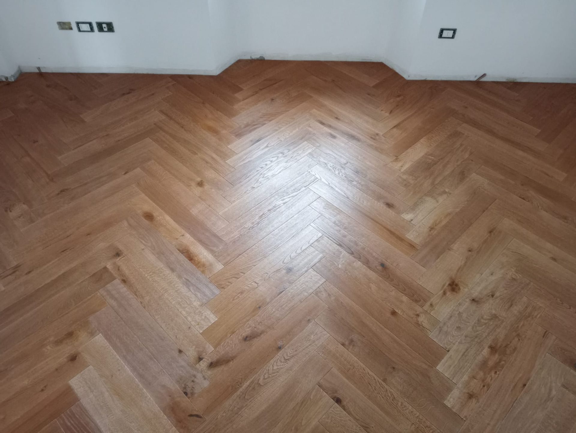 Pavimento in legno a spina di pesce in una stanza con pareti bianche. Il pavimento è di colore marrone chiaro e riflette la luce.