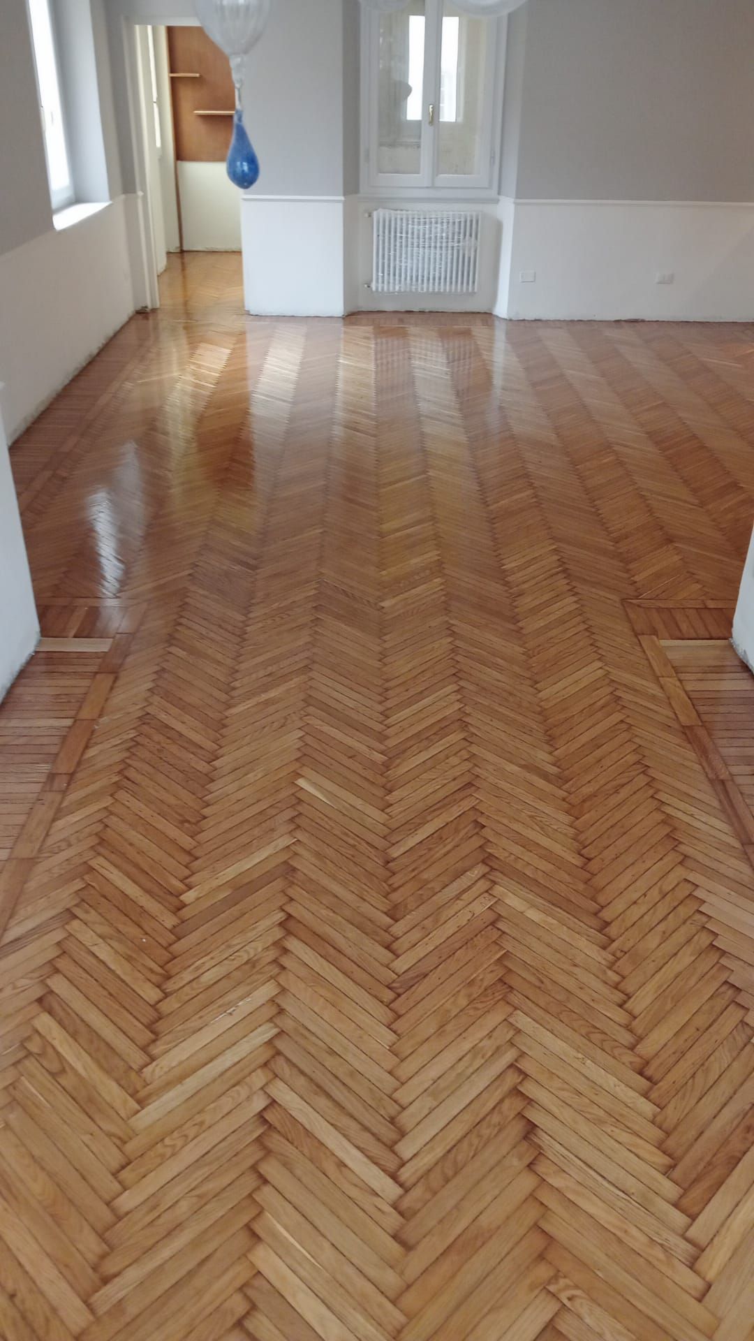 Pavimento in legno a spina di pesce in una stanza con pareti bianche, una porta e una finestra. Il pavimento è lucido e riflette la luce.