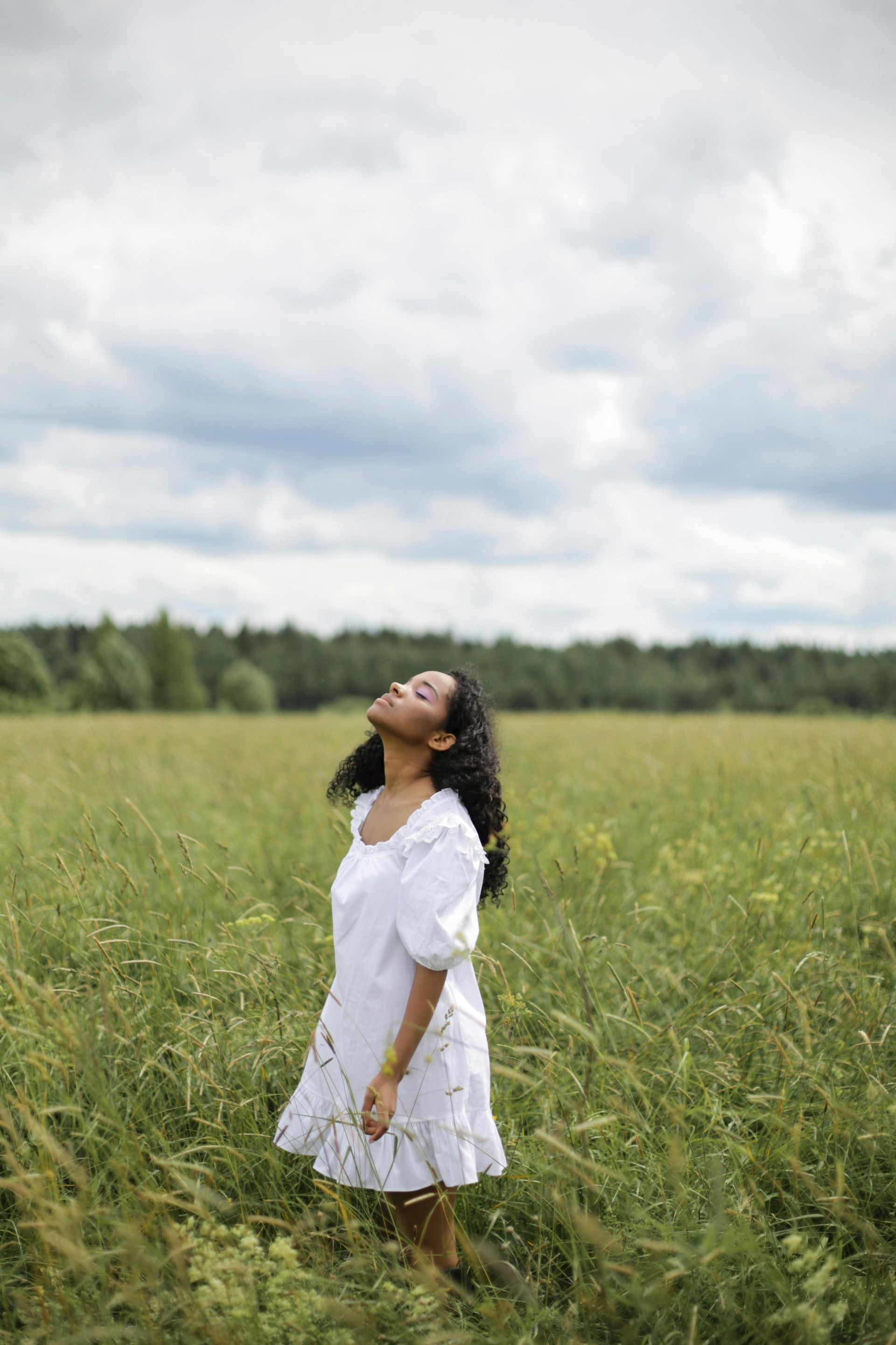 A woman in a white dress is standing in a field looking up at the sky.