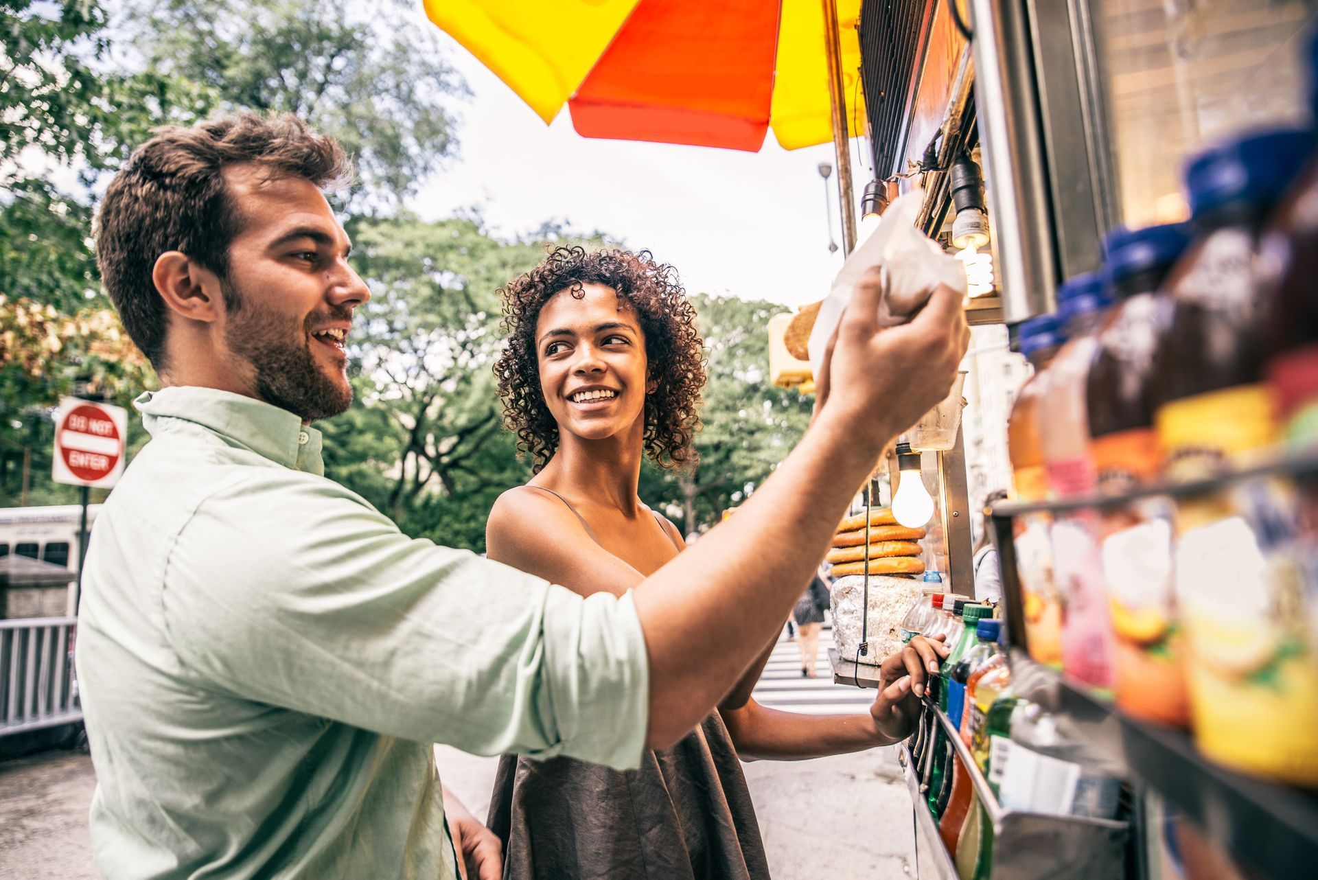 A smiling couple points at drinks in a street vendor's stand. The man wears a green shirt, and the woman has curly hair.