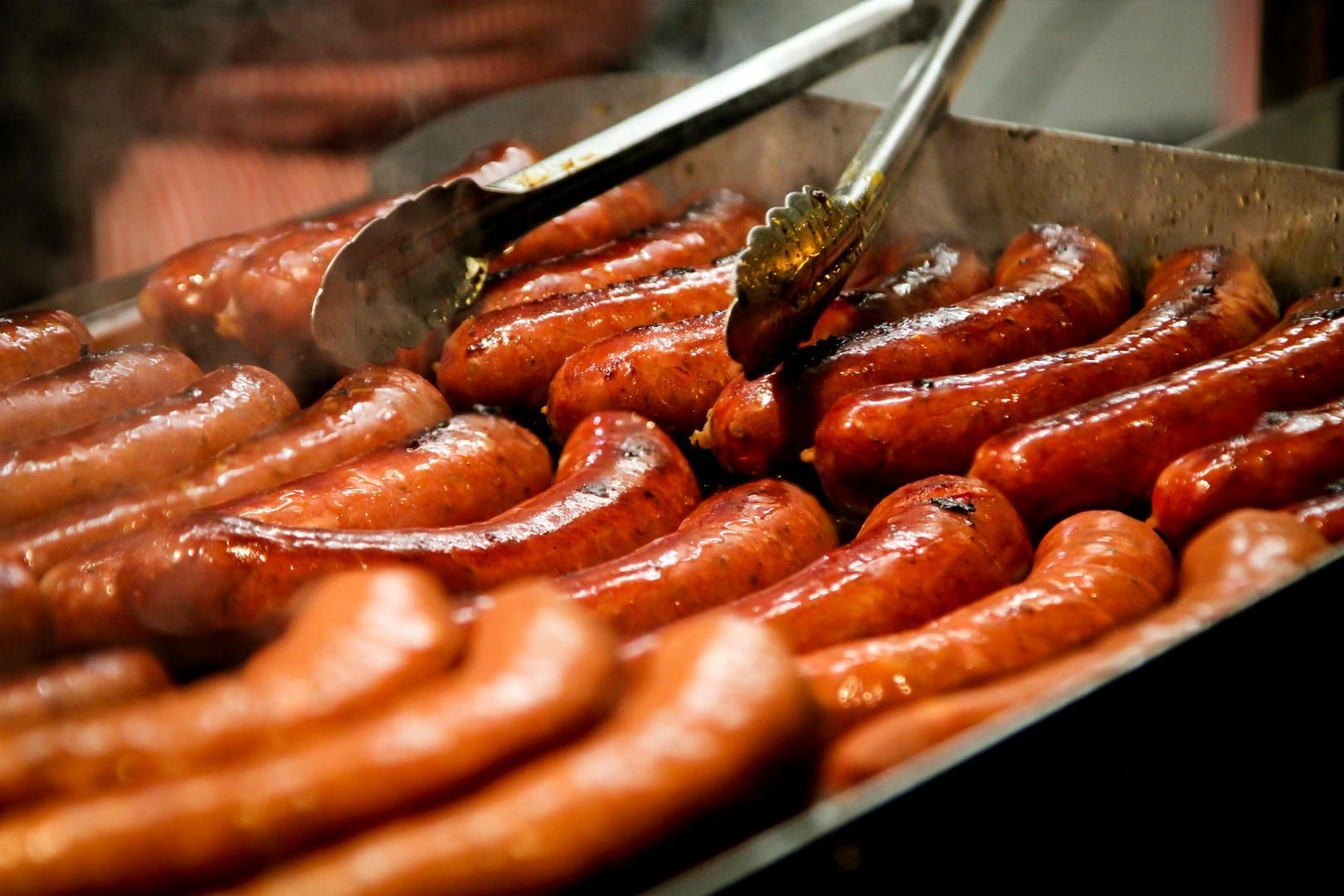 Grilled sausages on a hot griddle, being turned with tongs. The sausages are browned, with visible steam rising.