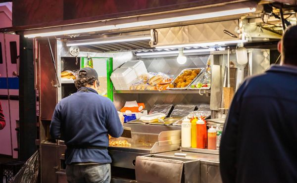 Two men stand at a lit food truck. One works at the counter, while another looks on. Food is displayed inside.