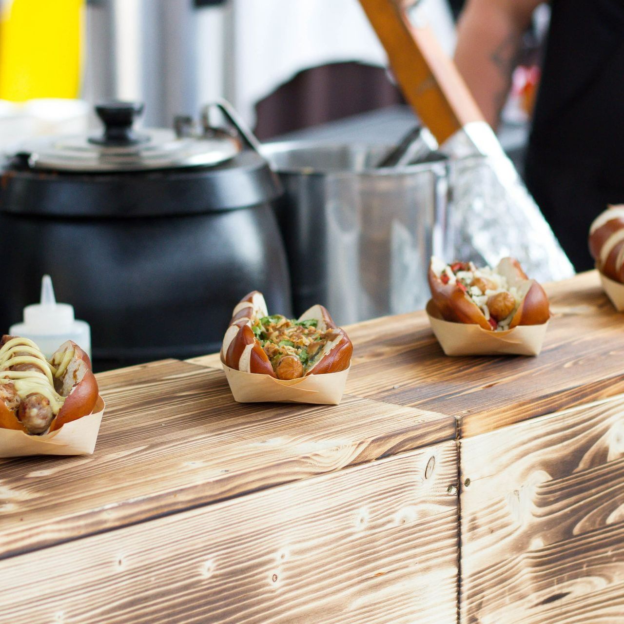 Hot dogs in pretzel buns on a wooden counter at a food stall, with toppings visible.