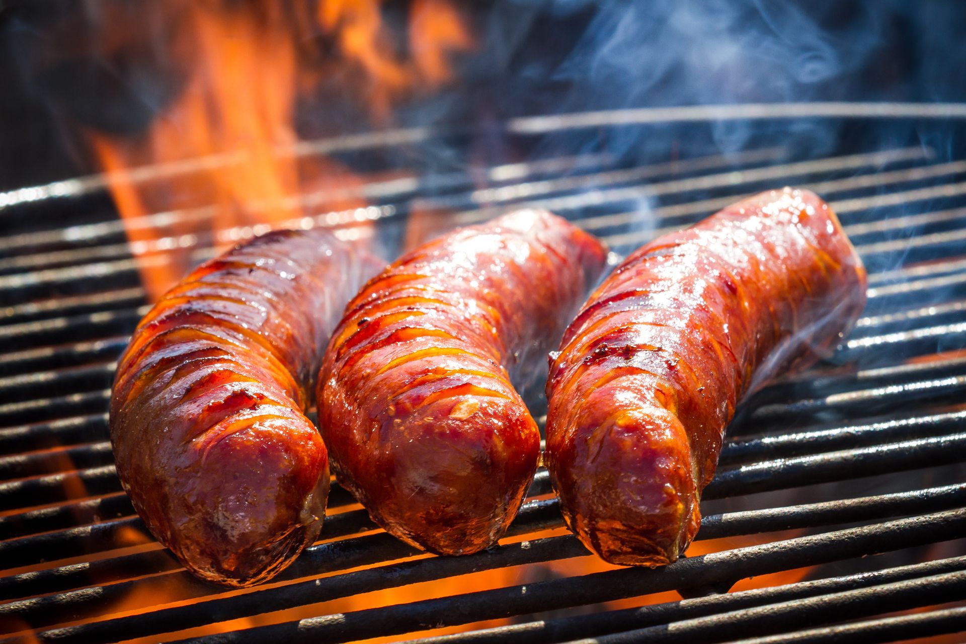 Three sausages grilling over an open flame, with smoke rising.