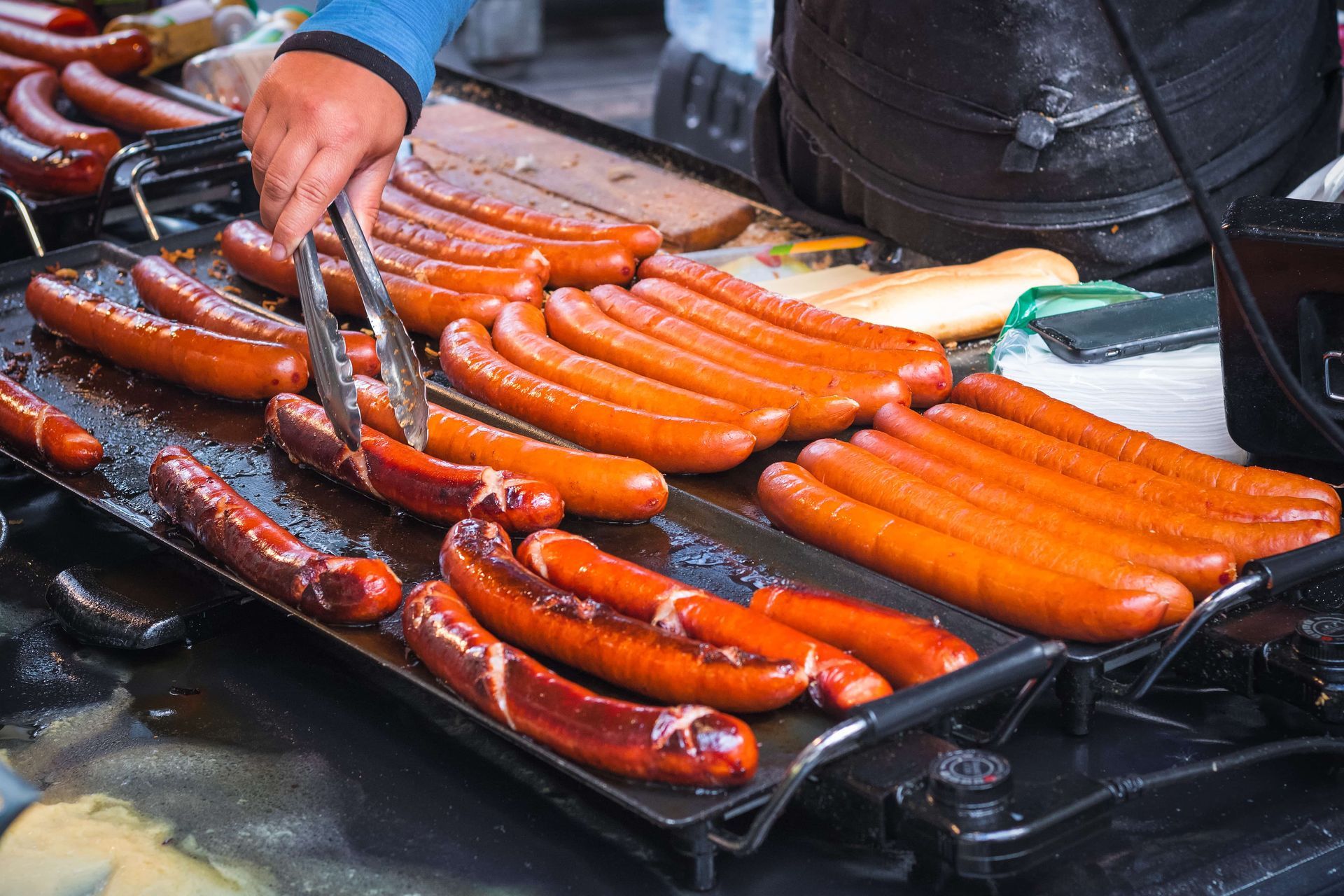 A person grilling rows of hot dogs on a flat griddle. The sausages are a reddish-brown color.
