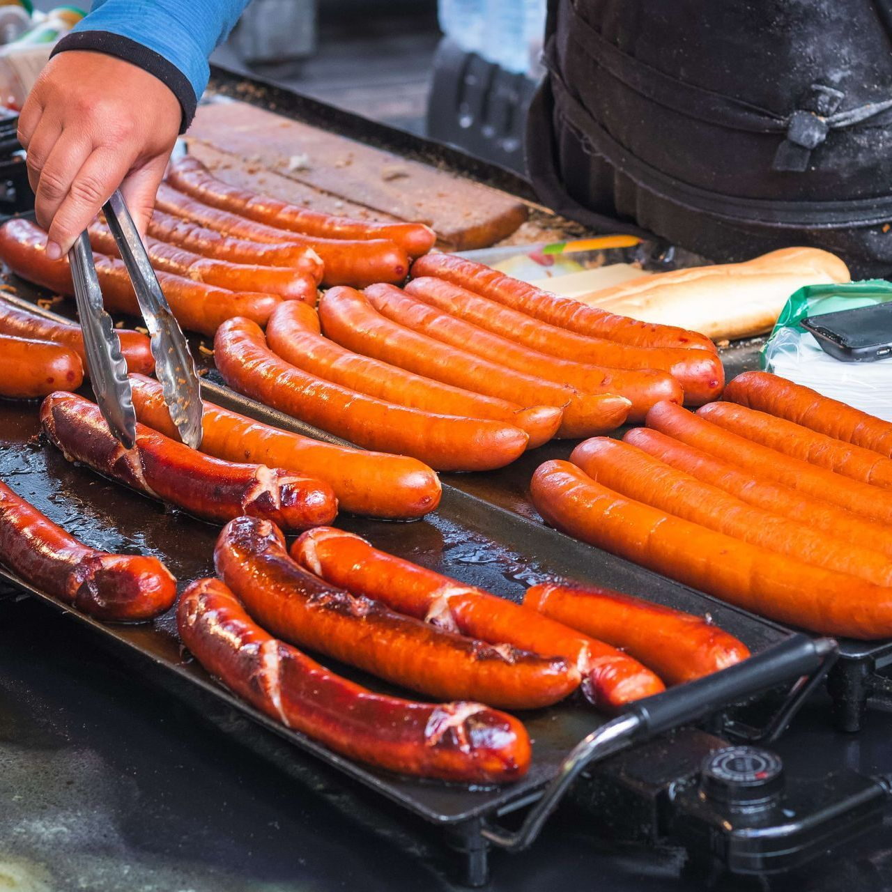 Sausages grilling on a black flat top grill at an outdoor food vendor; person flipping them with tongs.