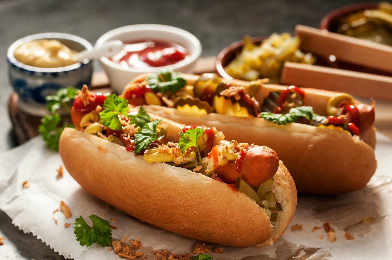 Two loaded hot dogs on a table, with bowls of condiments in the background. One is in focus, with mustard, ketchup, relish, and parsley.