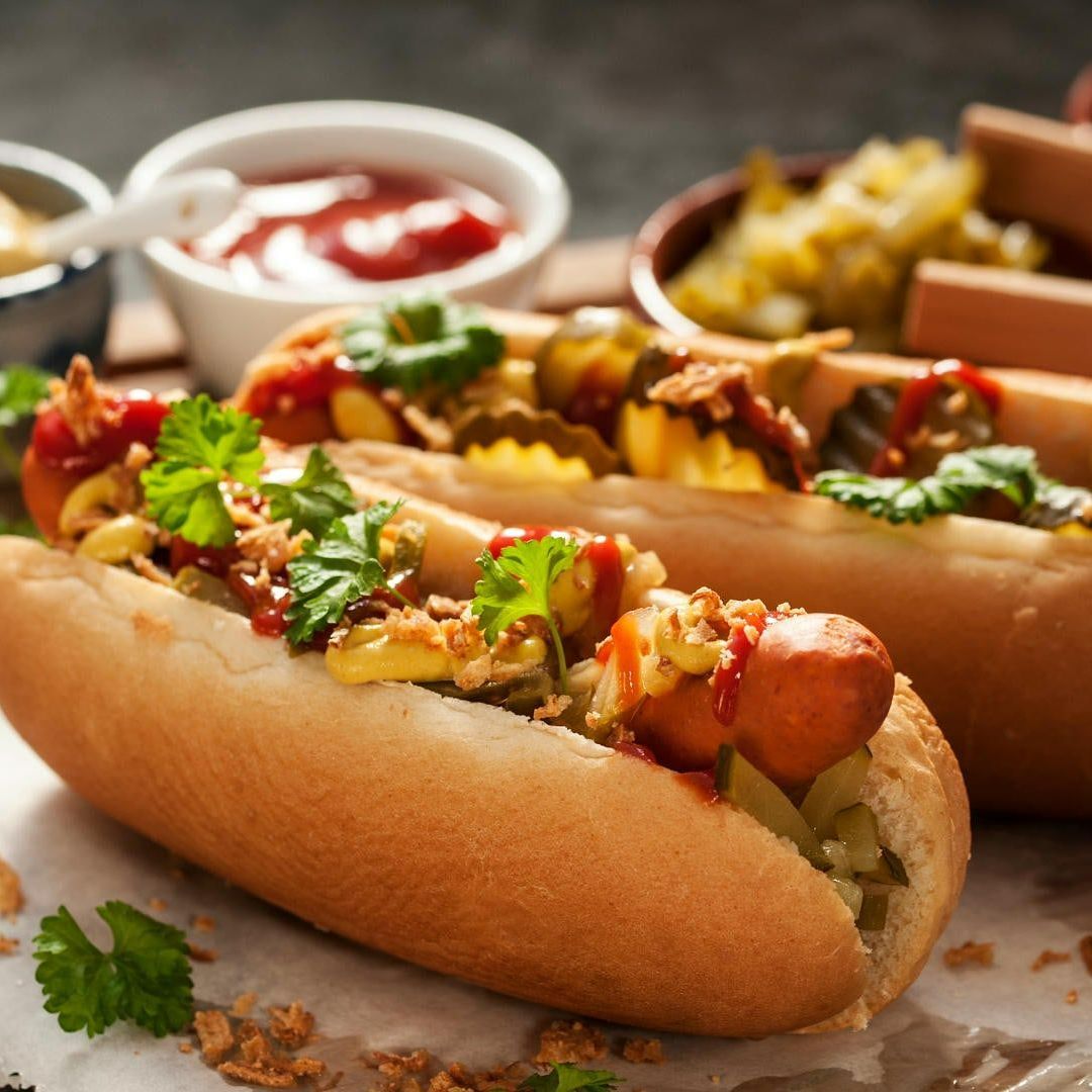 Two hot dogs in buns, topped with various condiments and garnished with parsley, alongside small bowls of ketchup, mustard, and relish.