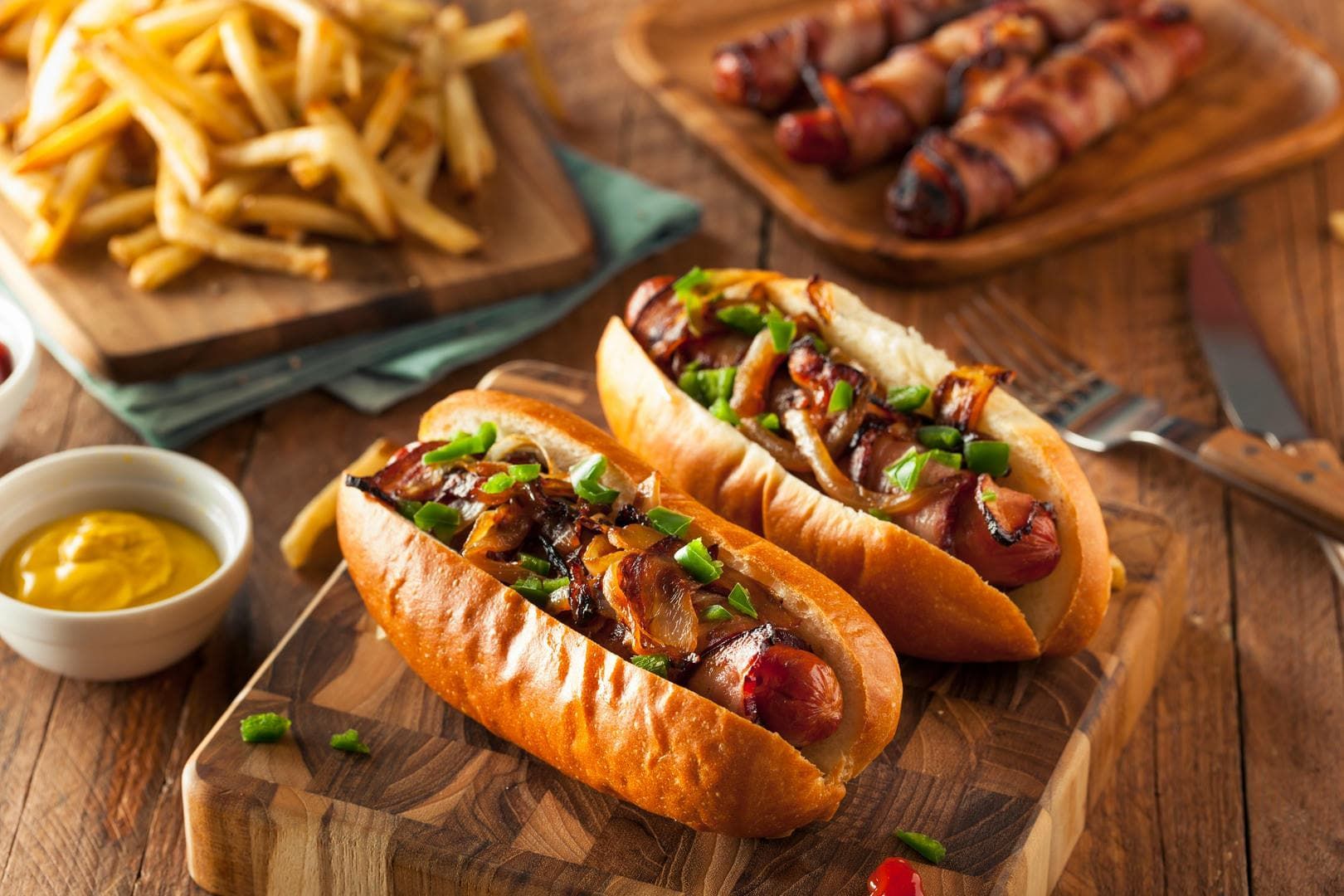 Two loaded hot dogs on buns with fries, mustard, and bacon-wrapped sausages on a wooden table.