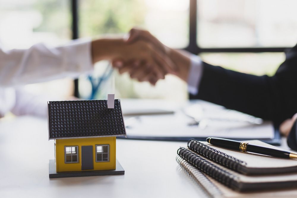 Two People Shake Hands Over a Miniature Yellow House on a Table — Into Finance Lending Solutions in Old Bar, NSW