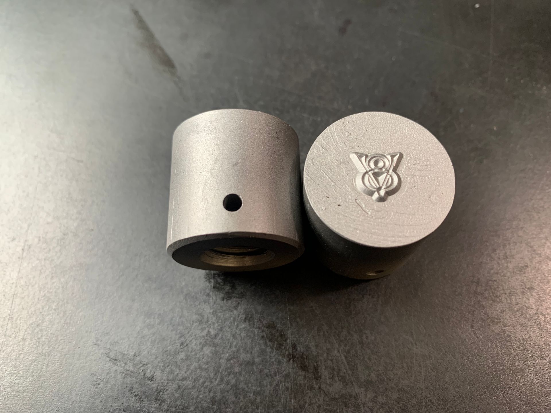 Two small, cylindrical metal objects; one with a hole, the other with an emblem.