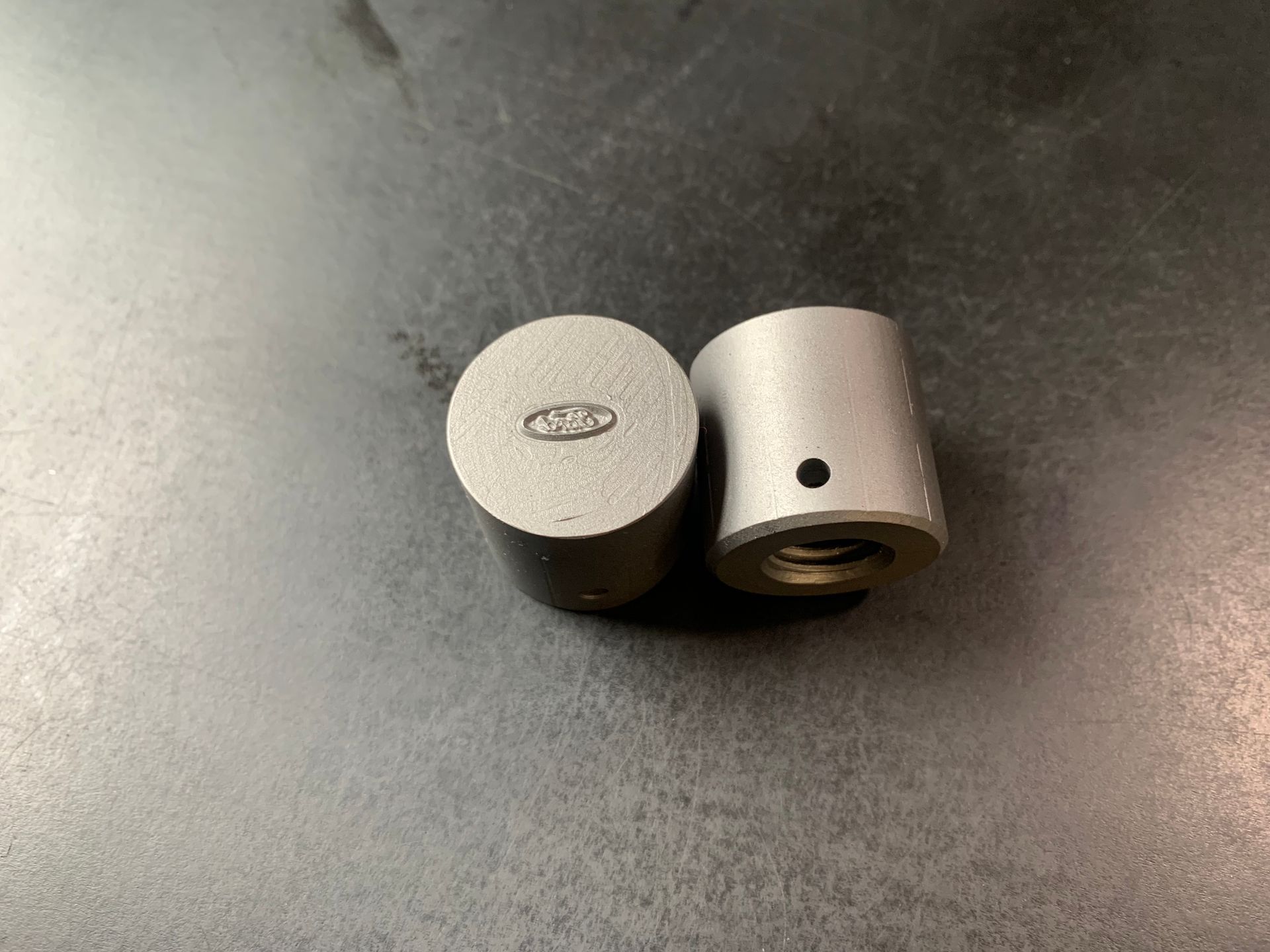 Two small, cylindrical metal objects with a threaded interior and a small hole. One has an oval marking.