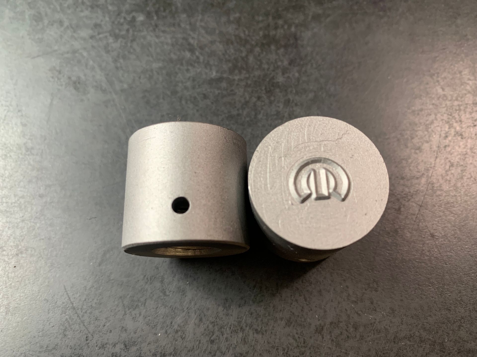 Two cylindrical metal pieces on a gray surface, one with a hole, the other with a logo.