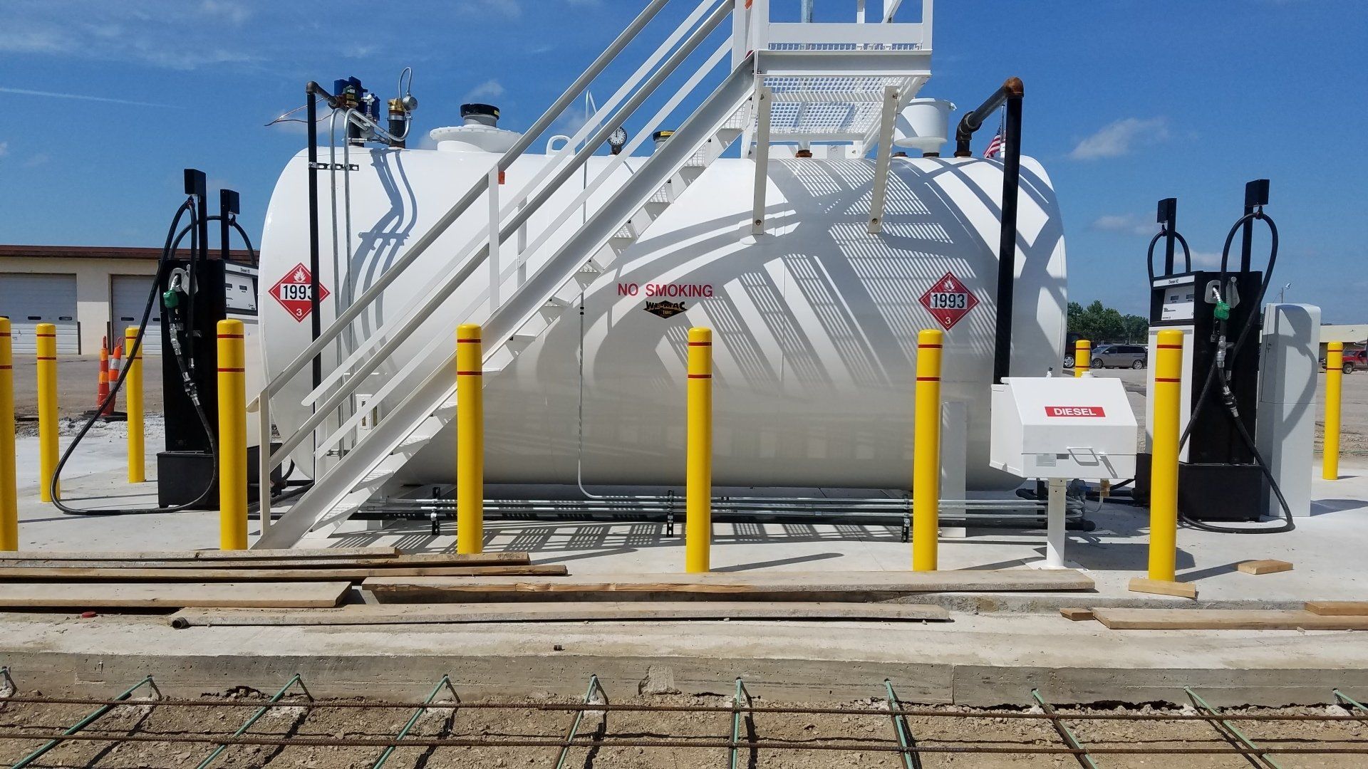 White fuel tank with yellow safety bollards, stairs, and black fuel pumps.