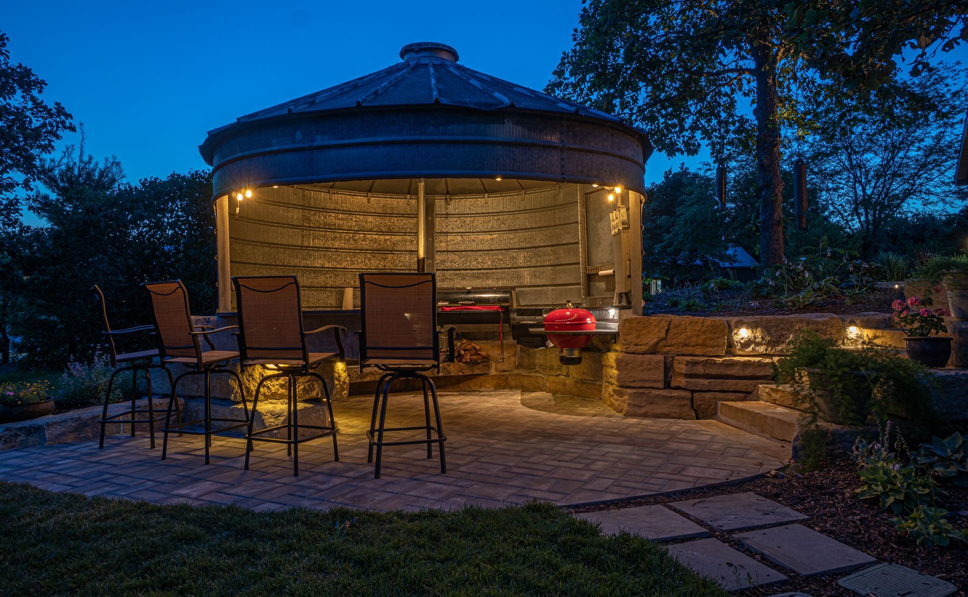 Modern covered patio with built-in grill, dining area, string lights, and surrounding garden.