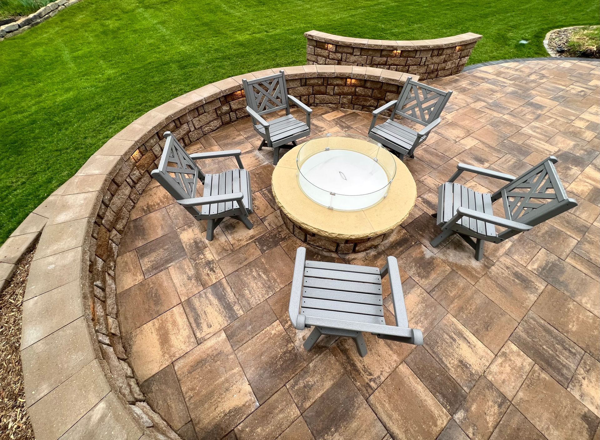 Stone patio with gray chairs around a circular fire pit, bordered by a low stone wall and green lawn.