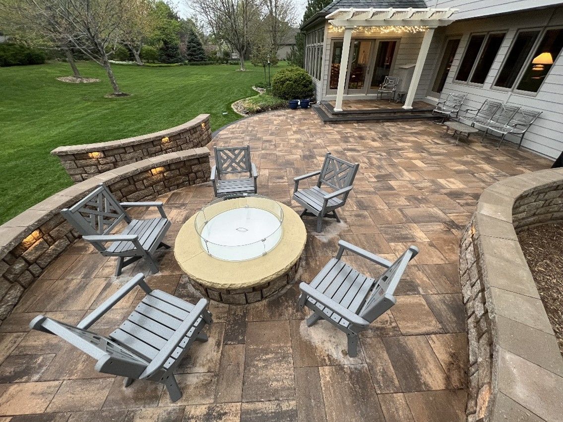 A spacious stone patio with five grey chairs surrounding a circular fire pit, enclosed by low retaining walls overlooking a green lawn
